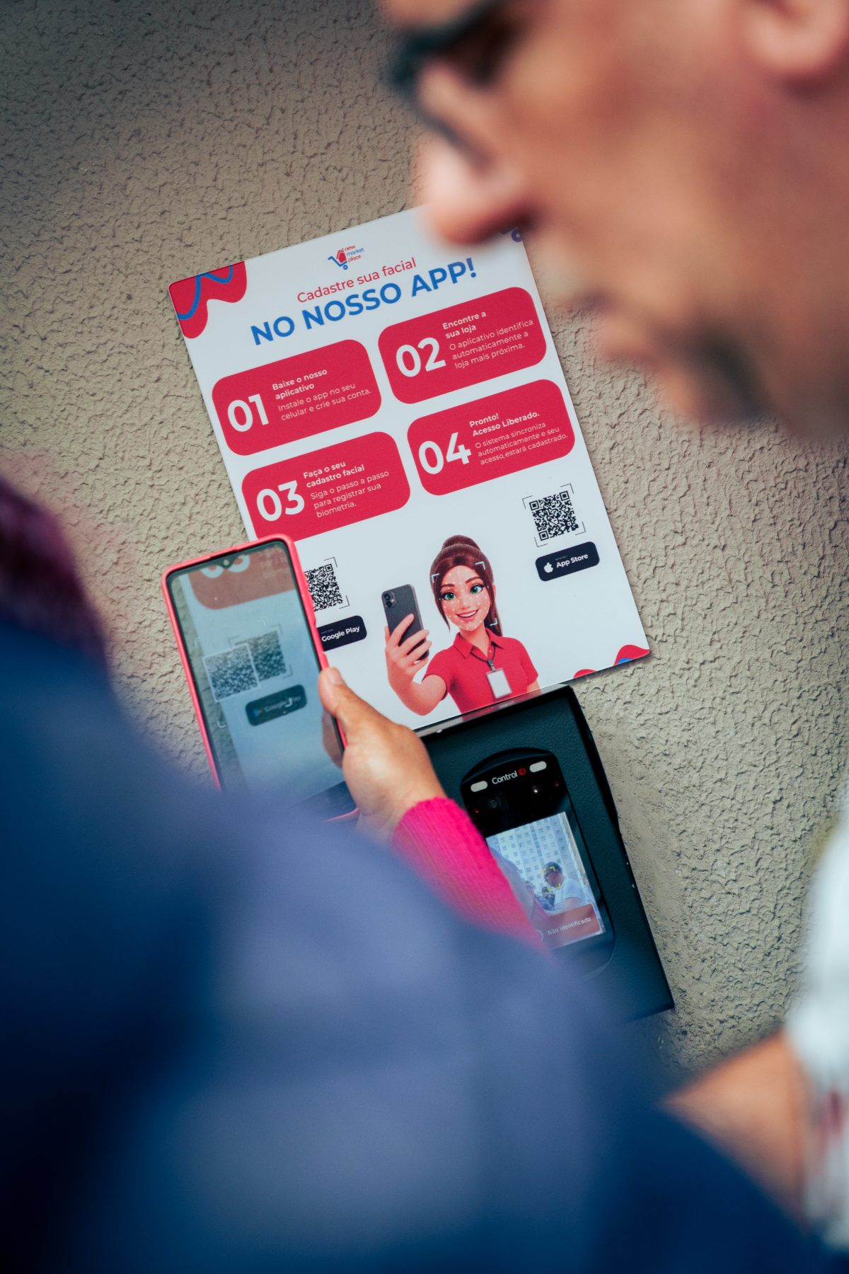 Morador aponta o celular para o QR code da placa “Cadastre sua facial no nosso app”, ao lado do leitor facial da porta