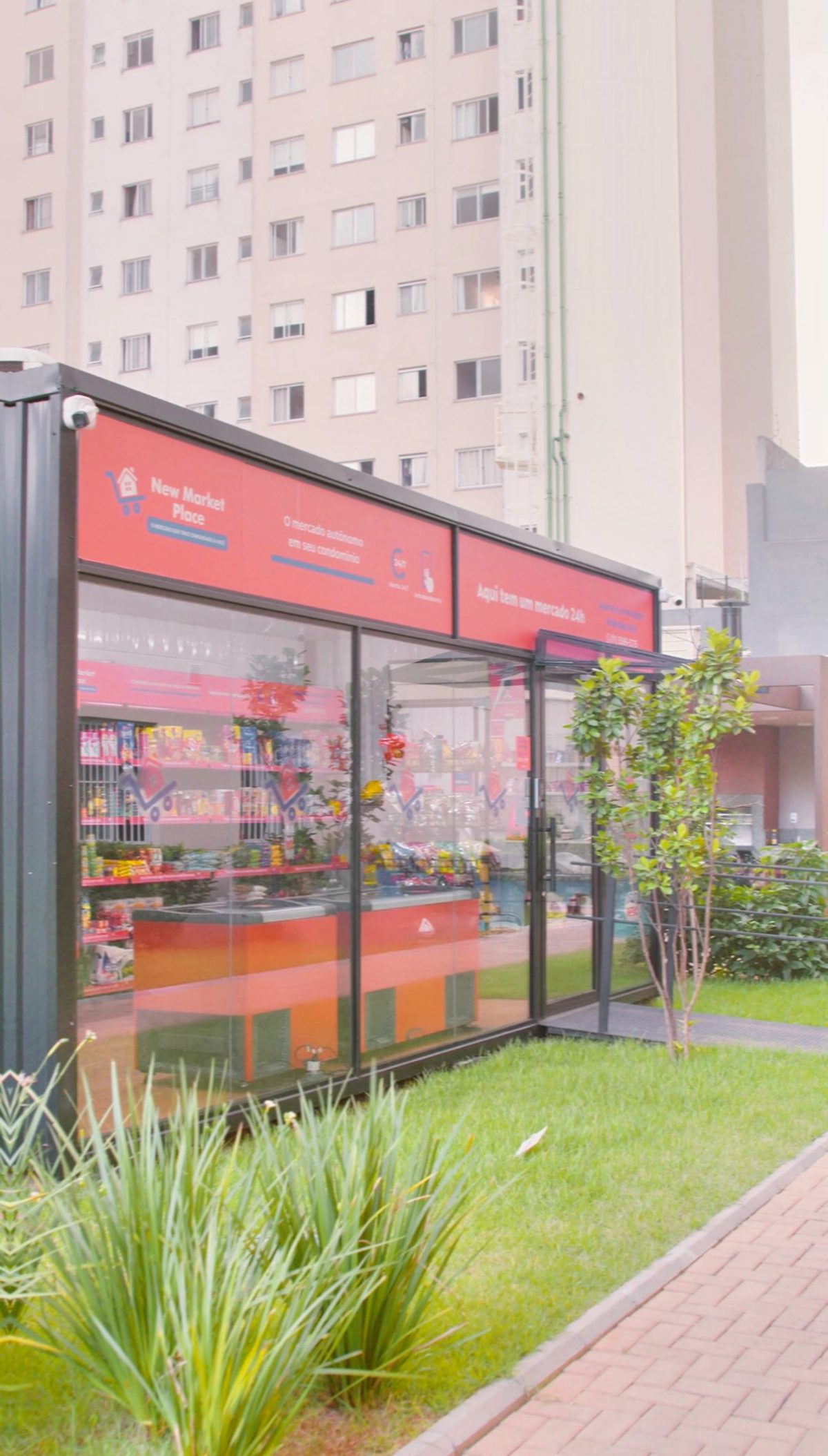 Unidade New Market Place em contêiner de vidro no jardim de um condomínio, com testeira vermelha e prédios ao fundo